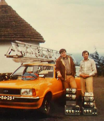 2 hams with pile of Drake and Heathkit gear standing beside car with tower on roof racks