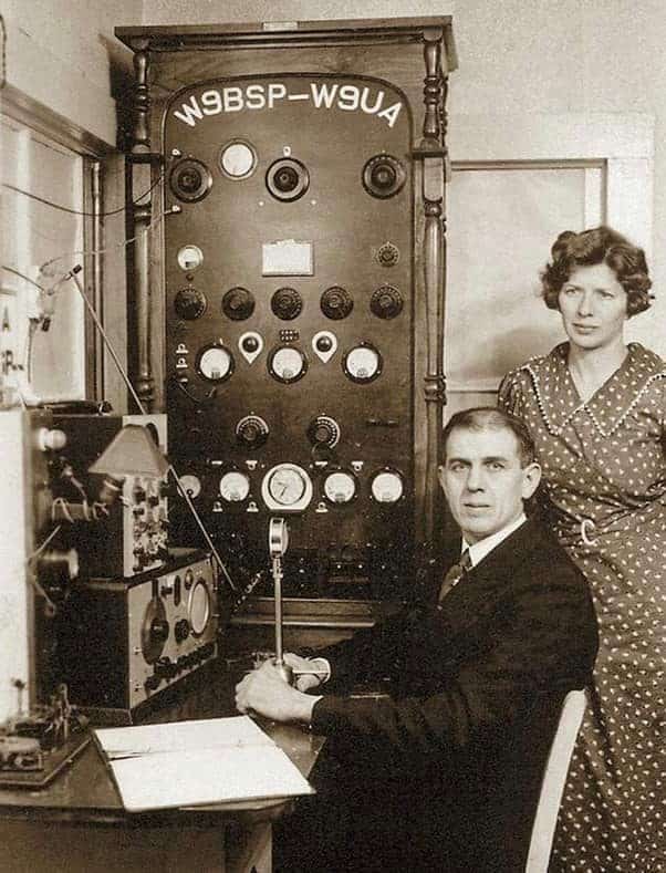 Man seated at desk of a 1937 hamshack with a microphone while woman standing behind him looks on. Two callsigns are displayed: W9BSP and W9UA.