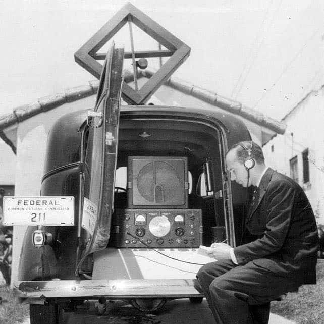 Rear view of an FCC station wagon with door open and man sitting on bumper listening to Hallicrafters receiver through headphones. Rotation directional aerial on roof.
