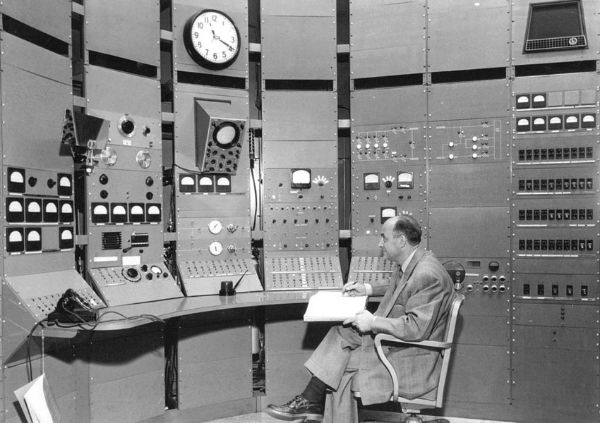 Man seated and taking notes in front of large control console