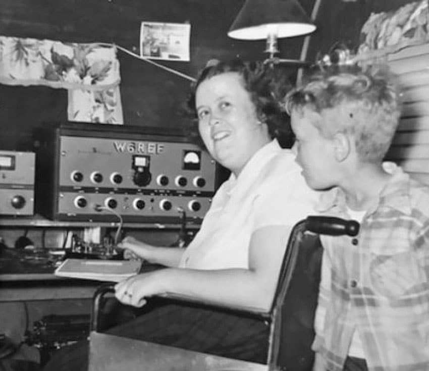 Betty Wilson W6REF seated in a wheelchair operating a bug ke in front of a Johnson Viking 1 transmitter. A young boy stands behind Betty.