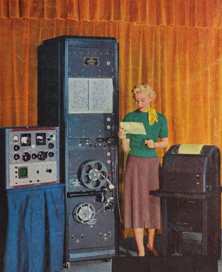 1956 publicity photo shows woman standing next to rack including a tapre recorder. On one side is a Super Pro 600 receiver and on the other is a teleprinter.