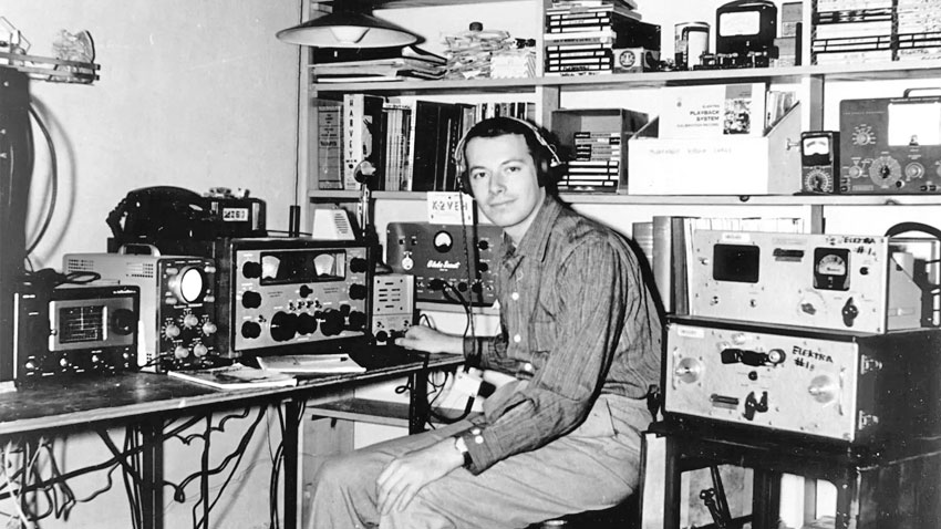 man wearing headphones seated in front of 1960s ham equipment. His hand may be resting on a morse key.