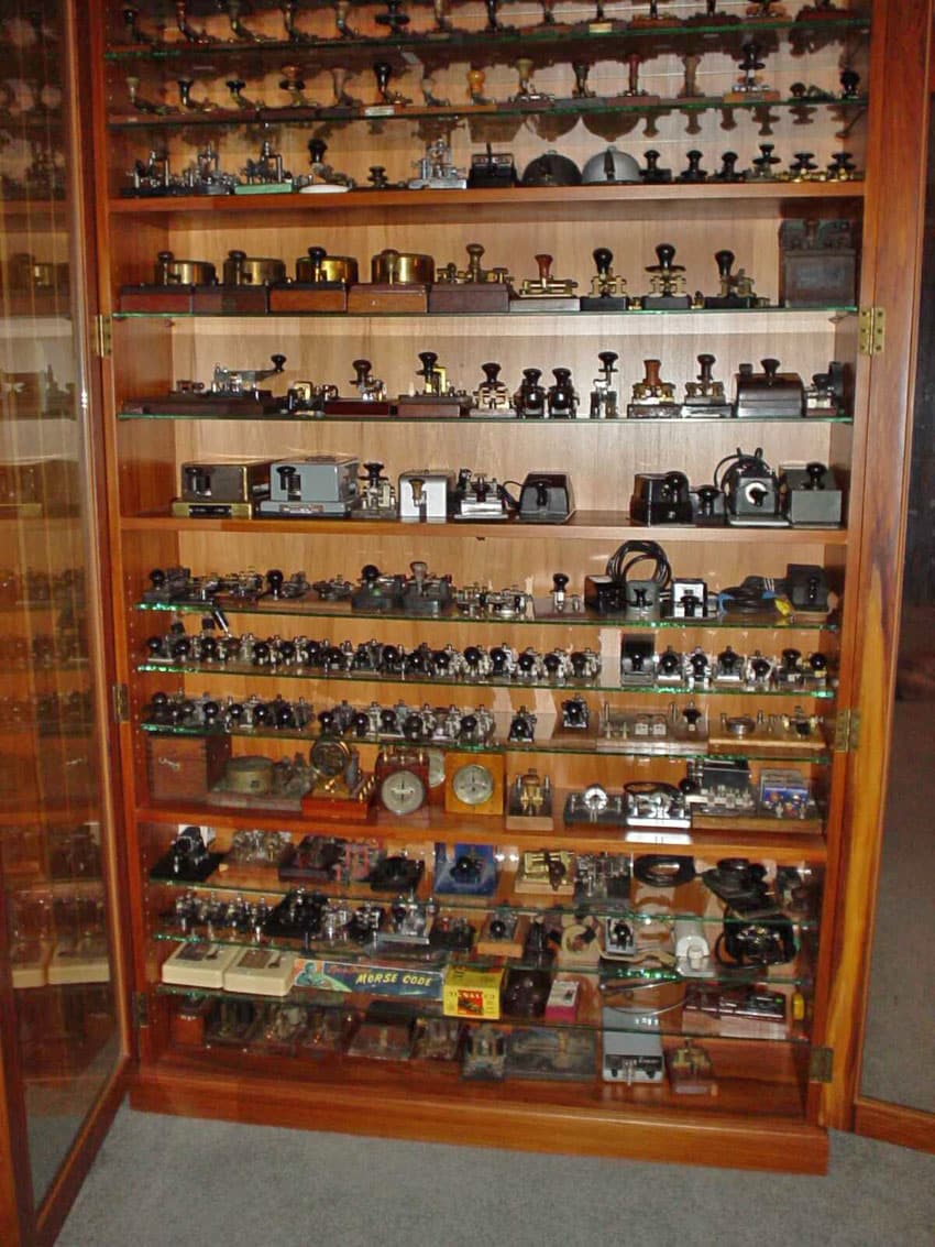 Wooden display cabinet with glass door containing Morse keys, mostly straight keys, but with a few bugs and paddles